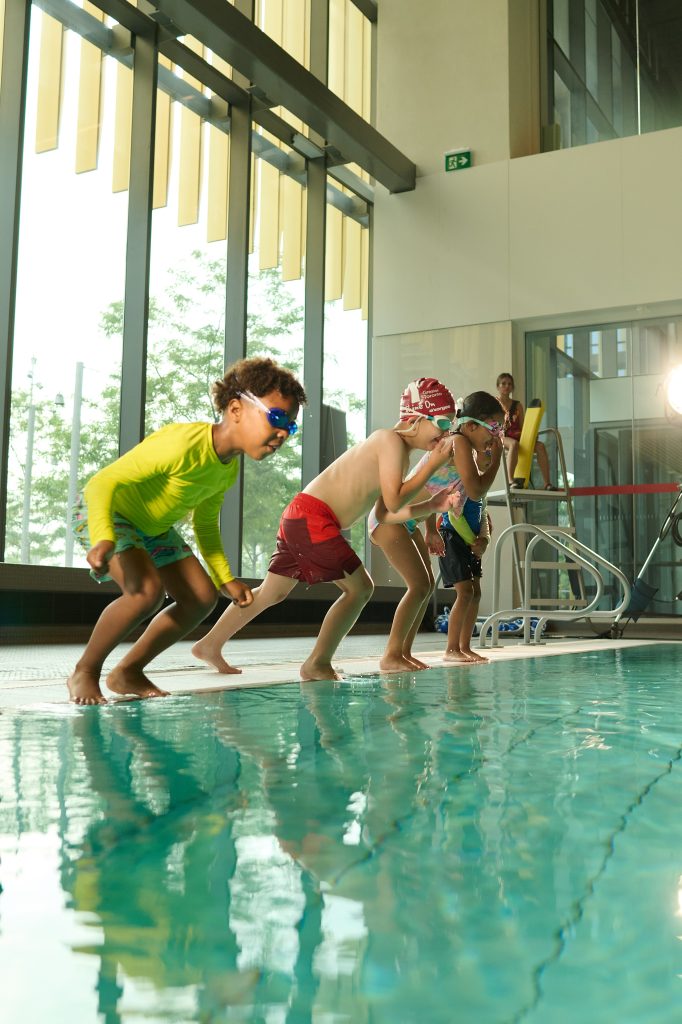 Kids in swim gear line up at pool edge, preparing to dive in an indoor swimming facility.